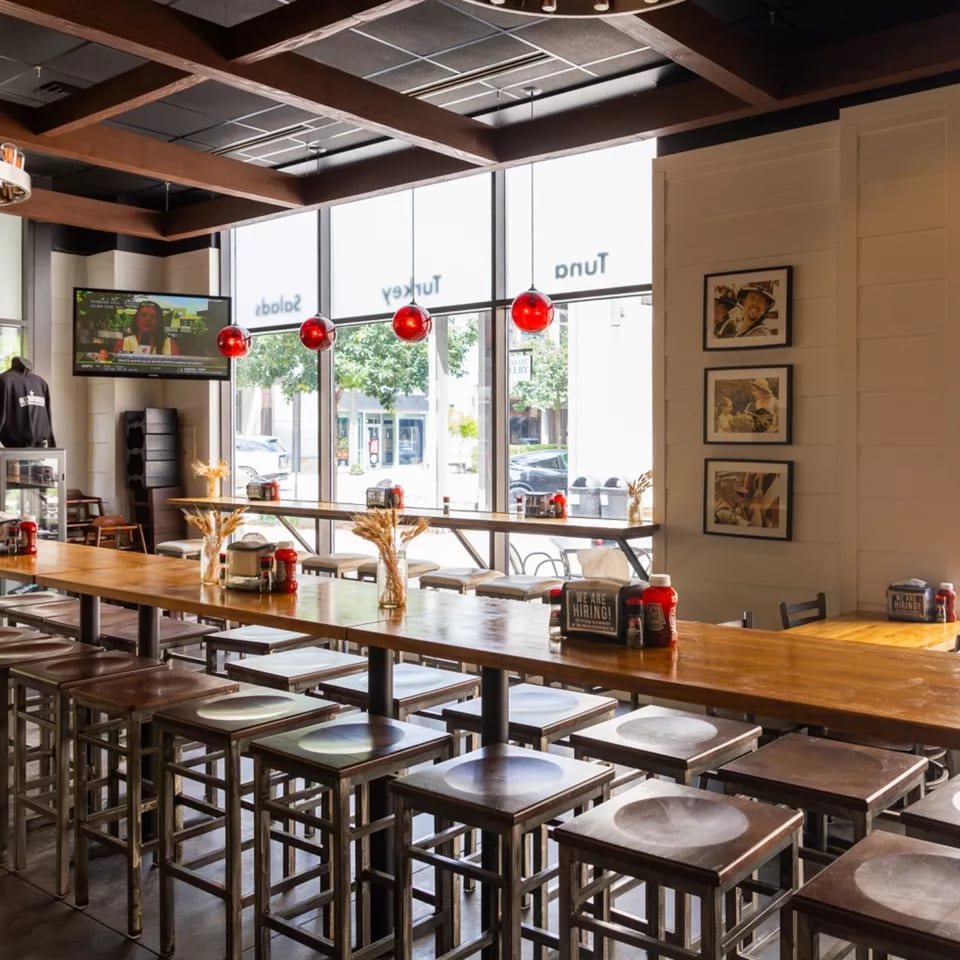 Interior of a modern restaurant with long wooden tables, stools, large windows, and red pendant lights.