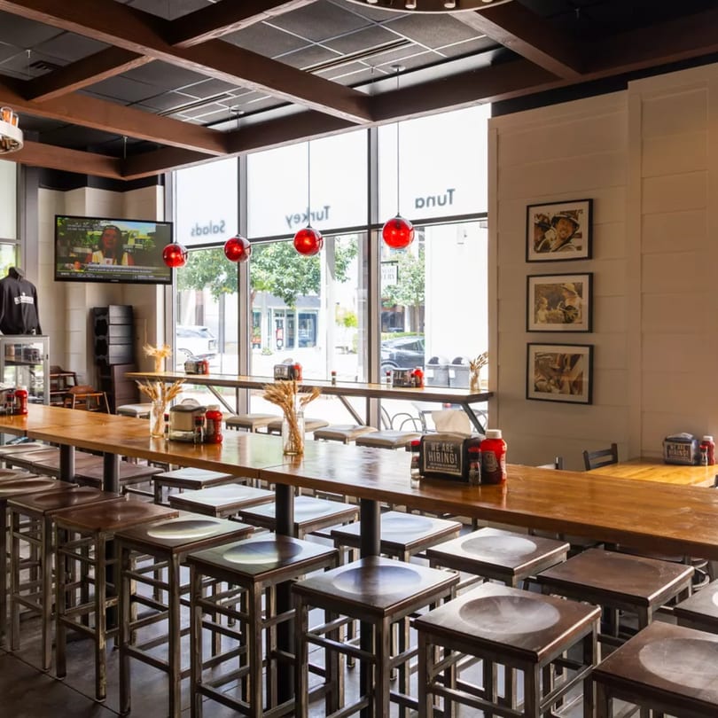 Interior of a modern restaurant with long wooden tables, stools, large windows, and red pendant lights.