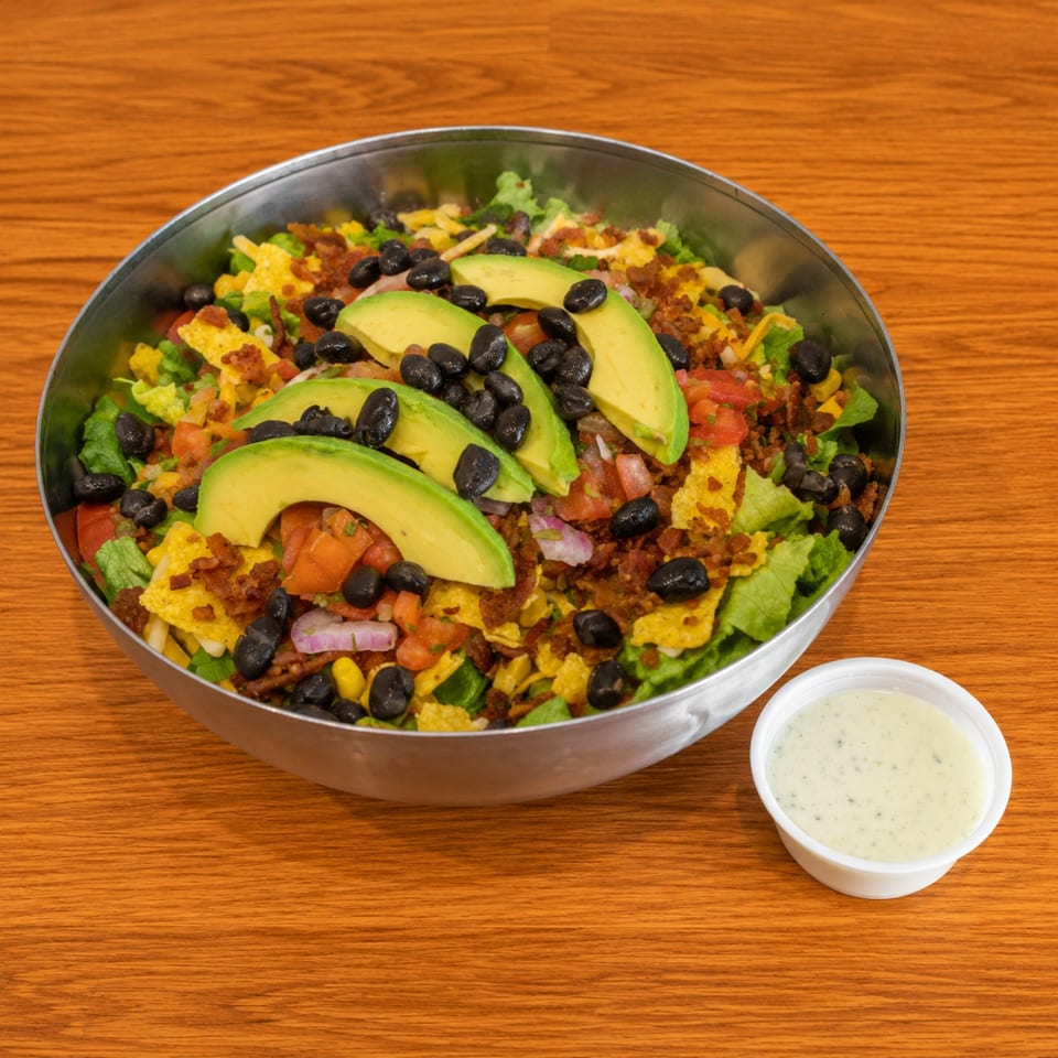 A large metal bowl of taco salad with avocado, black beans, and a side of dressing.