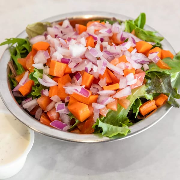 A fresh garden salad in a metal bowl, topped with diced red onions and carrots, with dressing on the side.
