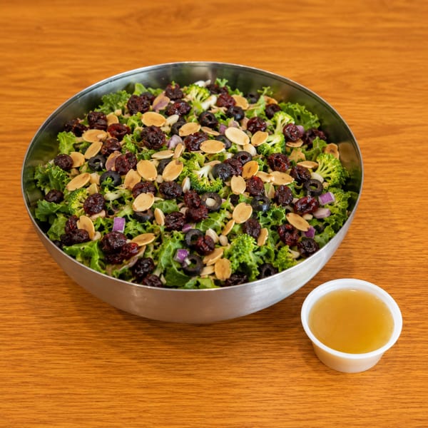 A large salad with kale, broccoli, dried cranberries, sliced almonds, and black olives, with dressing on the side.