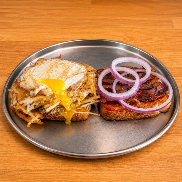 Open-faced sandwiches on a metal plate: one with hash browns and a fried egg, the other with bacon and red onion.